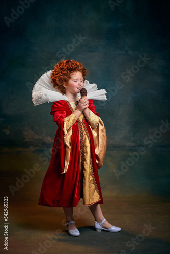 Portrait of little red-headed girl, child in costume of royal person licking ice-cream isolated over dark green background