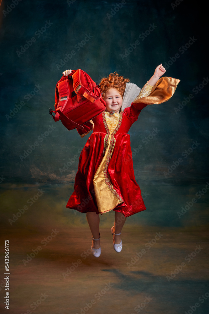 Fototapeta premium Portrait of little red-headed girl, child in costume of royal person jumping with school bag isolated over dark green background