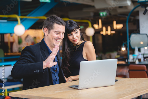 Smiling adult business man in formal clothing waving hand to someone during online video call using laptop while enjoying with wife in restaurant and office