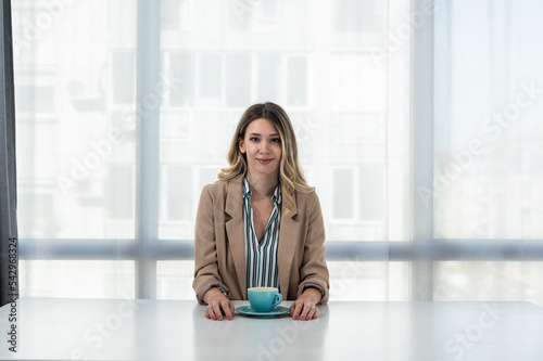 But first coffee. Young tired business woman in formal wear sitting in the office at empty desk with cup of coffee. Female white collar worker, freelancer financial expert morning routine.