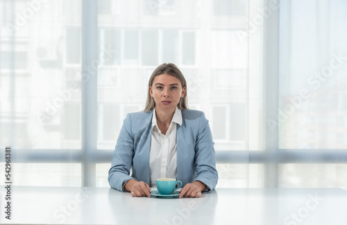 But first coffee. Young tired business woman in formal wear sitting in the office at empty desk with cup of coffee. Female white collar worker, freelancer financial expert morning routine.