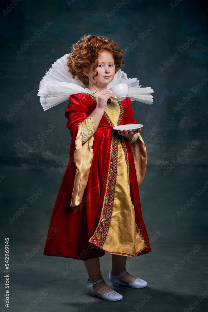 Portrait of little red-headed girl, child in image of royal person drinking tea isolated over dark green background
