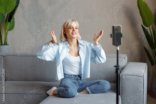 A young blonde girl is recording herself on the camera in the living room of her house, sitting on the couch. There is natural lighting, she is wearing a white T-shirt and a blue shirt.