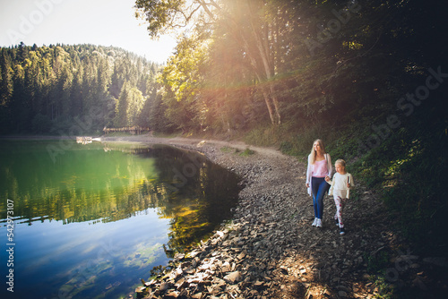 Mom and daughter walk hand in hand along the shore of a mountain lake on a bright sunny day. Coniferous forest around.