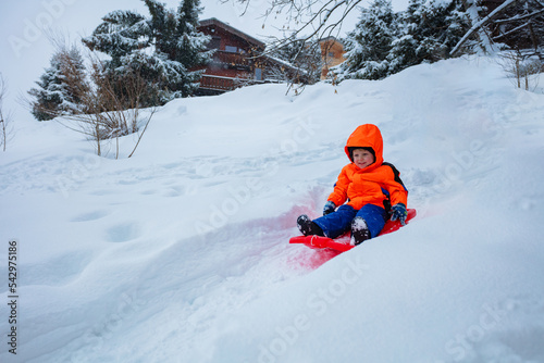 Happy boy in orange ski sport outfit go downhill on the sledge