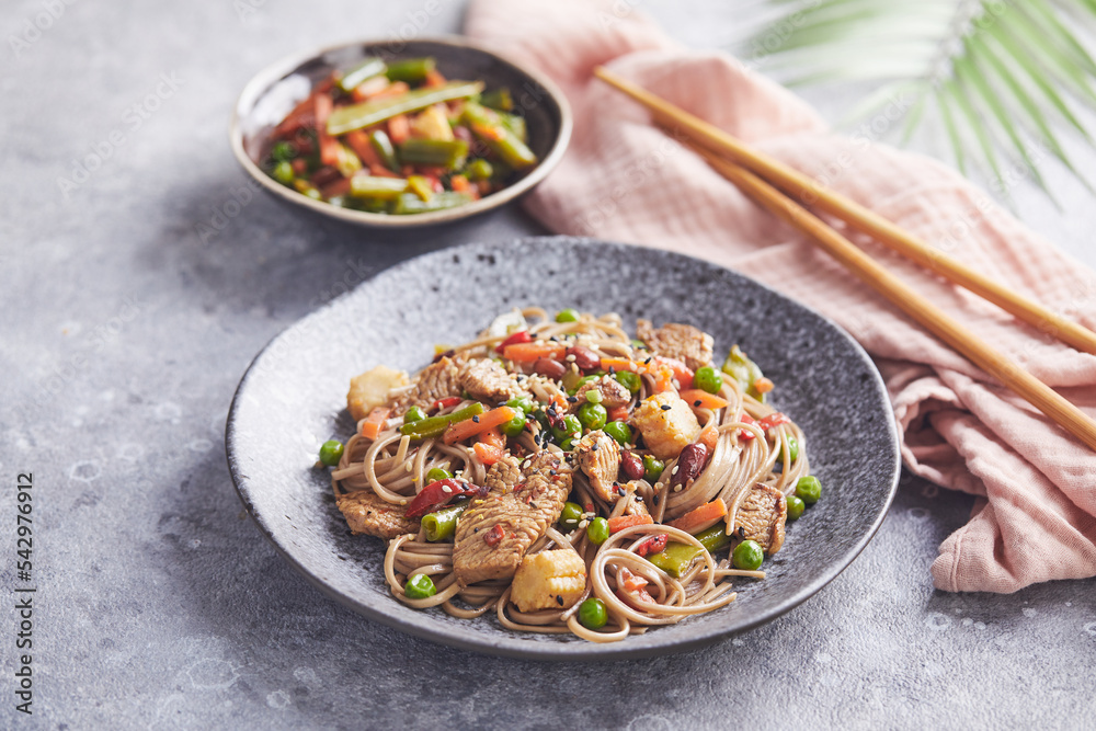 Wok with turkey meat, soba noodles, corn, green peas, green beans and carrots served on gray background with chopsticks. Asian food, concept of street food Wok with turkey meat, soba noodles, corn, green peas, green beans and carrots served on gray background with chopsticks. Asian food, concept of street food