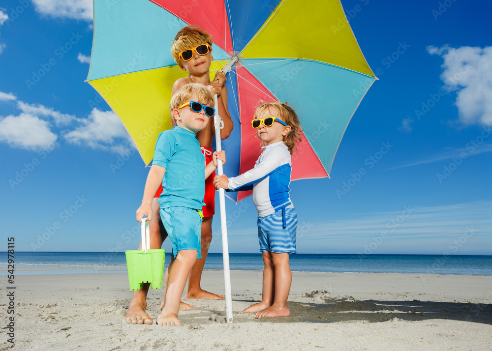 Three kids, boys, girl in sunglasses under parasol on a beach