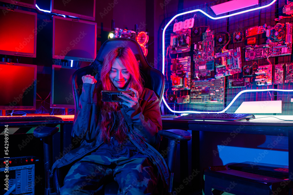 Fototapeta premium Winning. Happy Gamer playing video game online with smartphone with neon lights she raises hands to wins celebrating, confused Asian woman wearing gaming headphones feeling excited at home