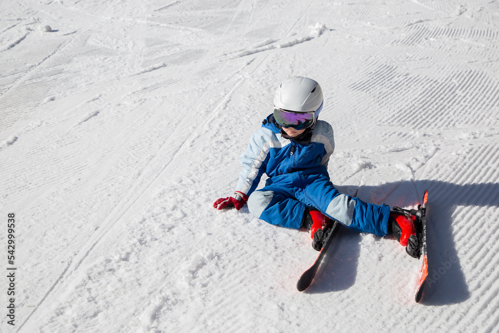 tired child in a helmet, ski goggles, skis and winter overalls sits on the snow. Fell into the snow, stopped to rest. winter fun. Little boy learning to ski during winter holidays on a sunny cold day