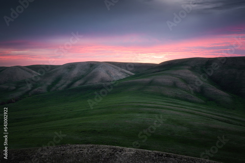 View of the hills and mountains covered by green grass under purple cloudy sky lightened by rising sun
