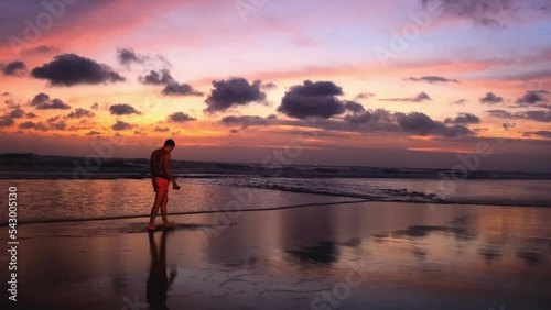 Full-length silhouette of man walking along ocean against backdrop of bright fiery multi-colored sunset. Man enjoying the peace and sound of the waves. Slow motion 4k footage of a beautiful landscape