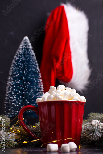 Red mug with hot cacao and heap of mashmellows, Santas hat and  blue decorative tree against black textured background. Christmas postcard. Place for text. photo