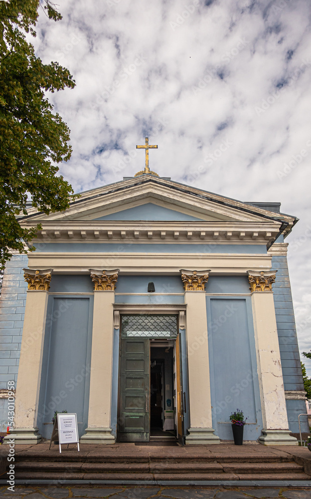 Finland, Hamina - July 18, 2022: Front entrance to blue St. Johns church, Johanneksen kirkko, under gray cloudscape. Decorated pillars, open door and cross on top. Some green foliage