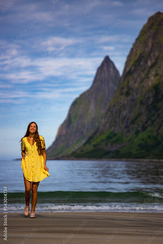 beautiful long-haired girl in a yellow dress walks on the famous beach with the mighty mountains in the background ersfjordstranda in norway, senja, holidays in the norwegian fjords