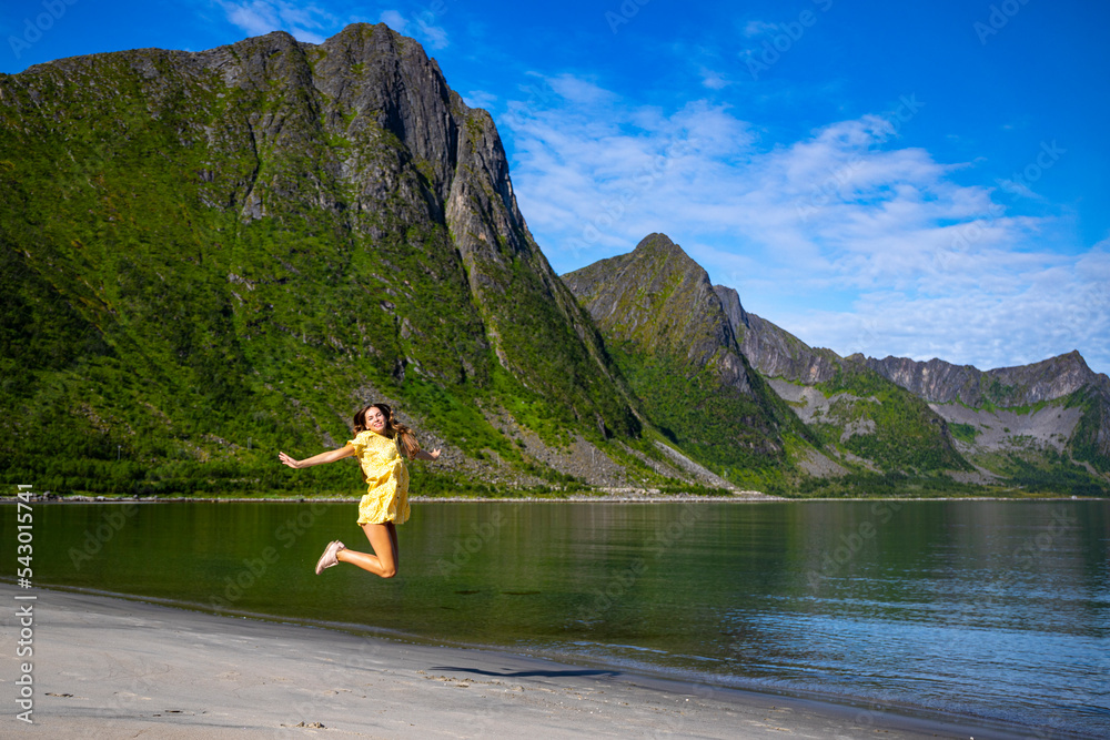 a beautiful girl in a yellow dress jumps for joy on a beach surrounded by mighty mountains on the island of senja in norway, holiday in the norwegian fjords, steinfjord