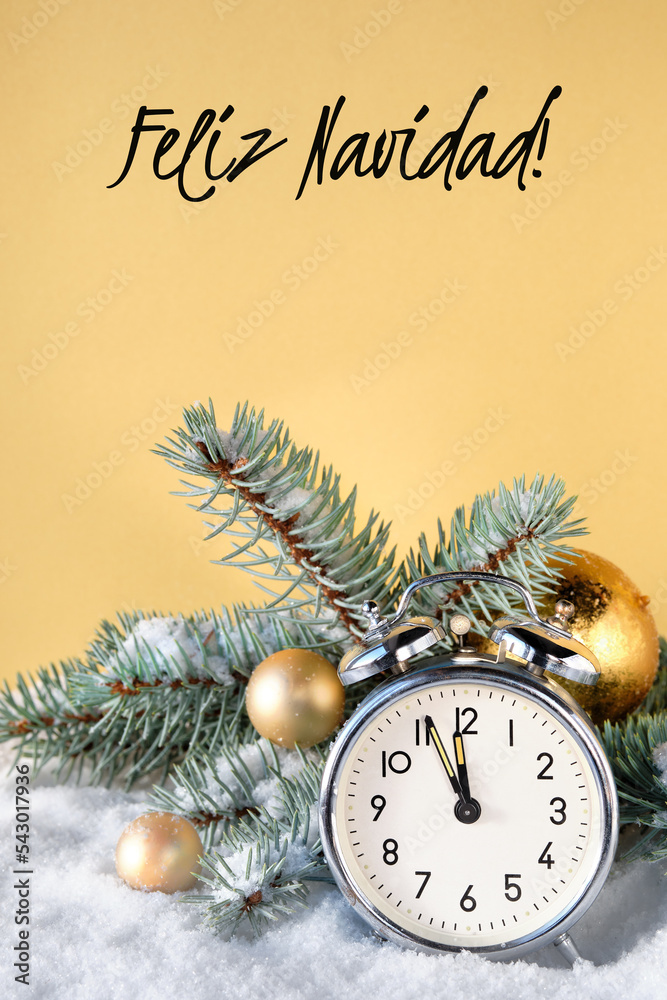 Old vintage alarm clock with fir twigs, gold trinkets and cones on snow.  Feliz Navidad meand Merry Christmas in Spanish language. Time ten to  midnight. Golden yellow background with text. foto de