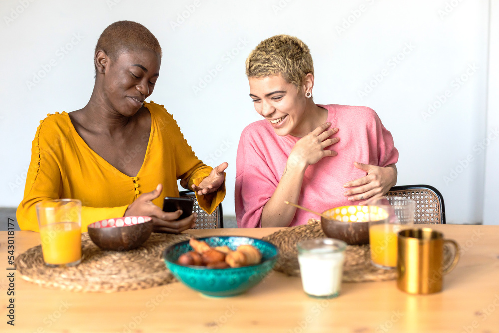 Horizontal shot of happy women discuss funny blog in internet, use mobile phone, eating healthy dish at home Horizontal shot of happy women discuss funny blog in internet, use mobile phone, eating healthy dish at home