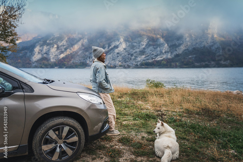 Obraz na płótnie Young woman traveler standing next to the car while traveling with her white swiss shepherd dog on the shore of a mountain lake w salonie
