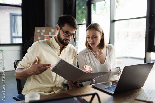 Businessman pointing at paper folder near intern with coffee and computer in office.