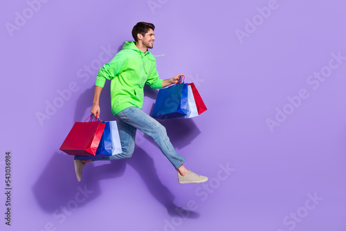 Full length photo of funky excited man wear green holding bargains jumping high empty space isolated purple color background