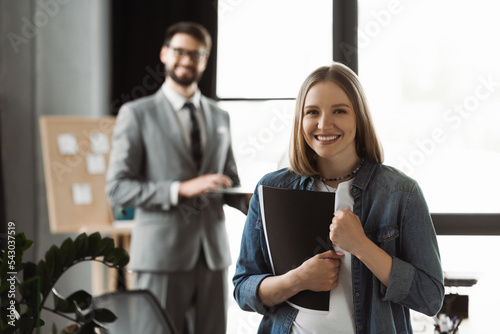Fotografija Cheerful woman with resume showing yes gesture near blurred businessman in office