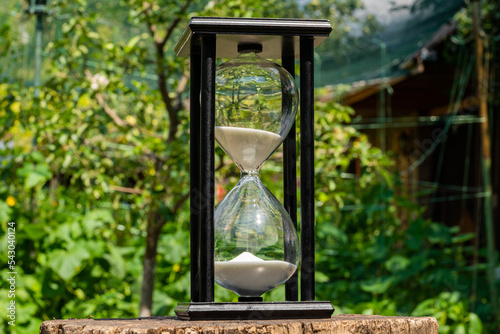 Old hourglass on wooden deck on a blurred background of a green garden. Glass sandglass in black wooden case with white flowing sand. Close up of timer counting time outdoors on a sunny summer day.