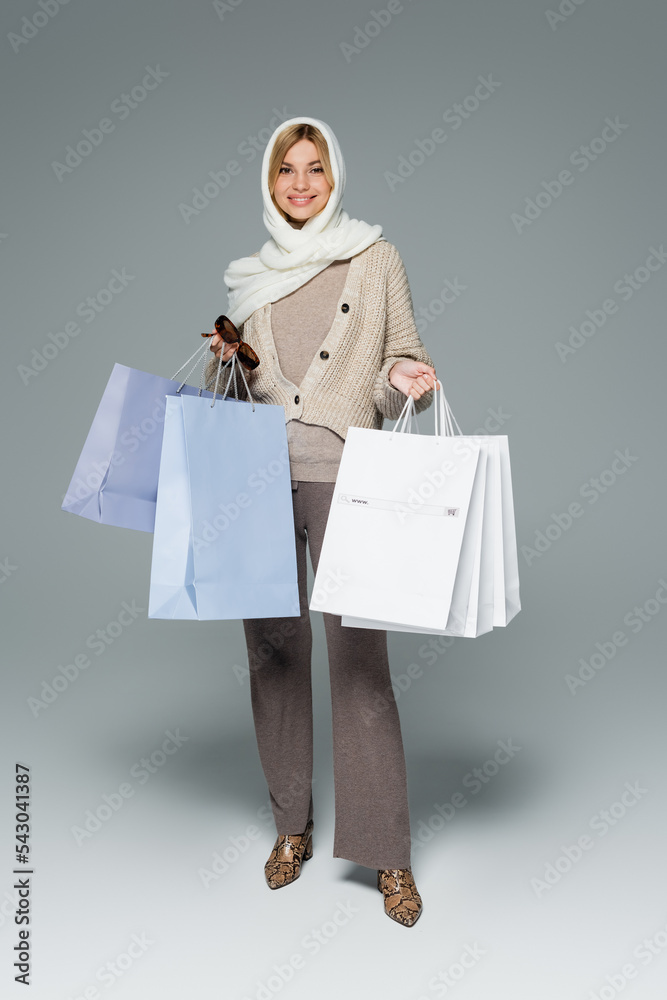 full length of joyful woman in winter headscarf holding stylish sunglasses and shopping bags on grey. full length of joyful woman in winter headscarf holding stylish sunglasses and shopping bags on grey.