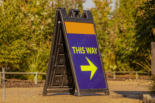 Room for Copy on Large  blue and orange standing plastic sandwich sign sitting on paved trail with blurred foliage in background with arrow saying This Way