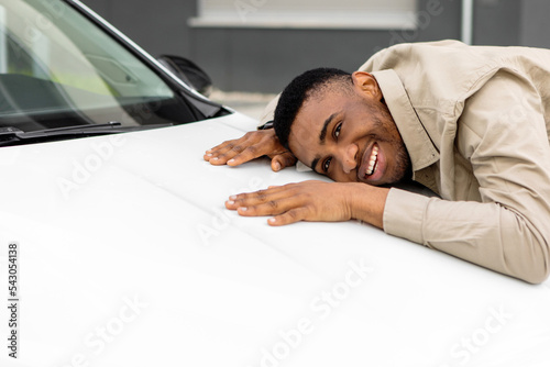 Happy african american man touching hugging his brand-new auto cherfully buying vehicle in auto dealership.