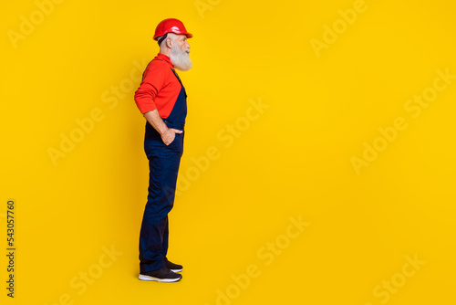 Photo of cheerful positive senior guy dressed uniform overall red hardhat arms hands pockets empty space isolated yellow color background © deagreez
