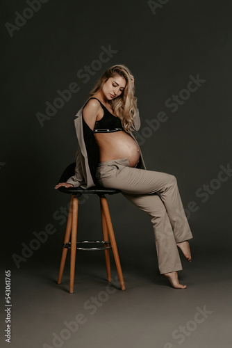 Front view of stylish pregnant woman posing in dark studio. Attractive girl with curly hair, dressed in a beige jacket and pants, leaning on chair in modern studio
