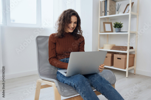 Side view of cheerful smiling happy curly beautiful lady enjoy online work using laptop doing NFT design project loves her job sitting in chair at home. Modern Profession and Remote Job