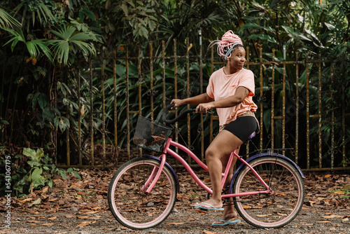 Imagen horizontal del perfil de una joven mujer afroamericana con cabello rosado en exterior dando un paseo en bicicleta en un día de verano.  photo