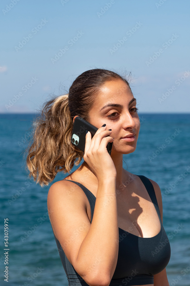 Malia, Crete, Greece. 2022. Portrait of an ttractive woman with ponytail hair using a mobile phone at the seaside. Malia, Crete, Greece. 2022. Portrait of an ttractive woman with ponytail hair using a mobile phone at the seaside.