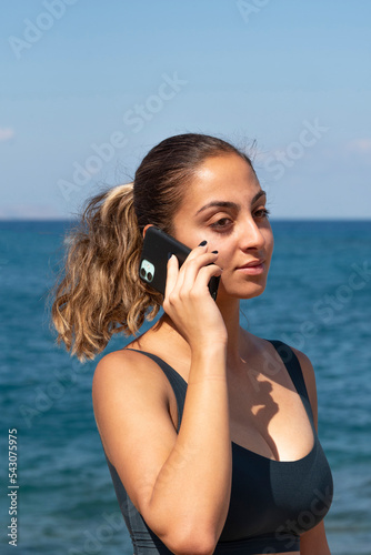 Malia, Crete, Greece. 2022. Portrait of an ttractive woman with ponytail hair using a mobile phone at the seaside.