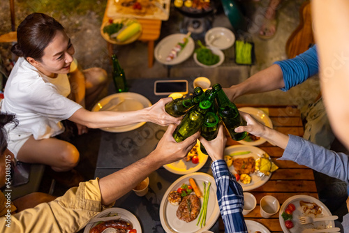 Group of Asian man and woman friends enjoy and fun outdoor lifestyle celebrating holiday event with toasting beer bottle and have dinner together near the tent while camping on summer travel vacation.