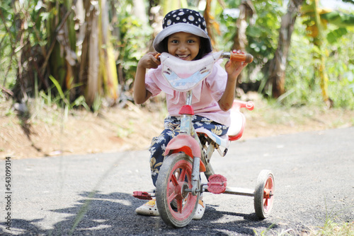 daytime portrait, a little girl wearing a floral hat smiling happily while pushing a tricycle