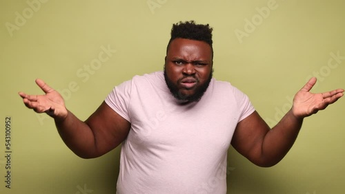 Annoyed frustrated bearded African-american man wearing pink t-shirt standing with raised hands and indignant face asking why. Indoor studio shot isolated on light green background.