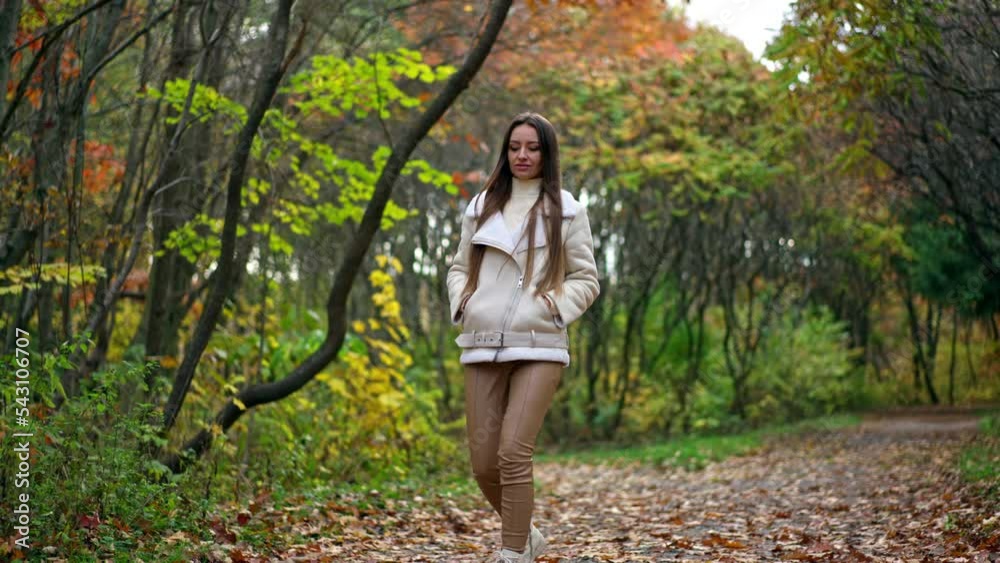 Relaxed long-haired brunette tossing fallen leaves walking by the road. Calming leisurely stride in the autumn park. Relaxed long-haired brunette tossing fallen leaves walking by the road. Calming leisurely stride in the autumn park.