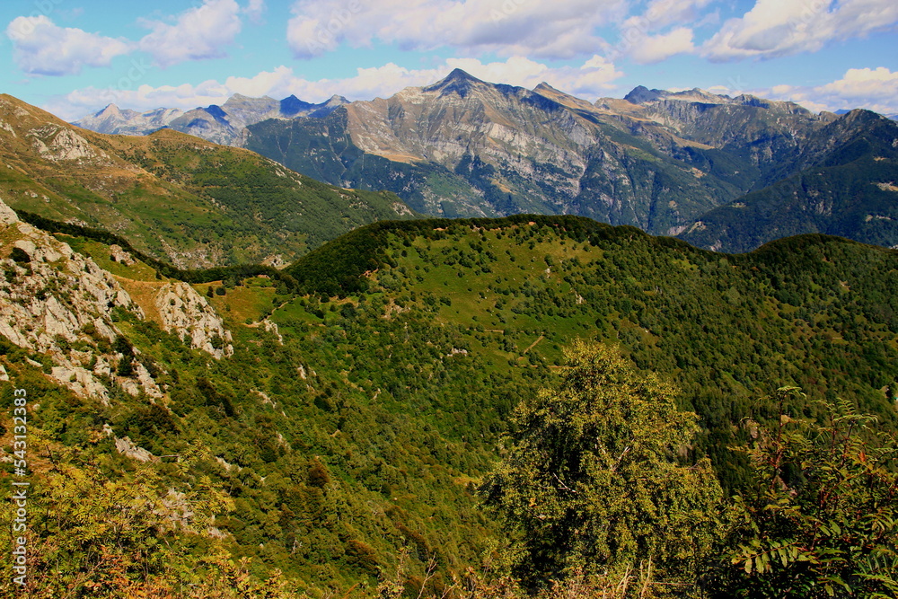 Panoramic view of the landscape with mountains and green hills in the foreground against a blue sky with clouds on Mount Cimetta, near Locarno in Switzerland Panoramic view of the landscape with mountains and green hills in the foreground against a blue sky with clouds on Mount Cimetta, near Locarno in Switzerland