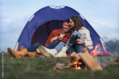 A happy couple is sitting and talking in front of a tent by the fire while enjoying the sunset. They enjoy camping in nature.
