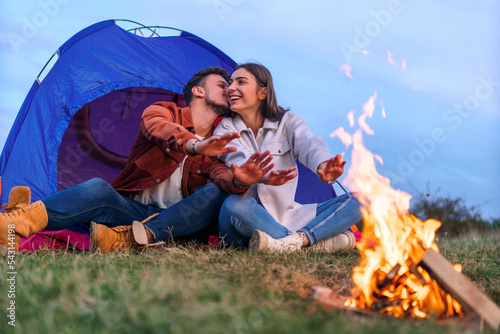 A happy couple is warming their hands and camping by the fire. Behind them is a blue tent.