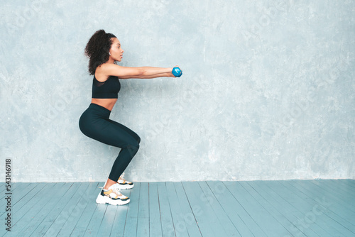 Fitness smiling black woman in sports clothing with afro curls hairstyle.She doing squats. Young beautiful model with perfect tanned body. Female holding dumbbells in studio near gray wall