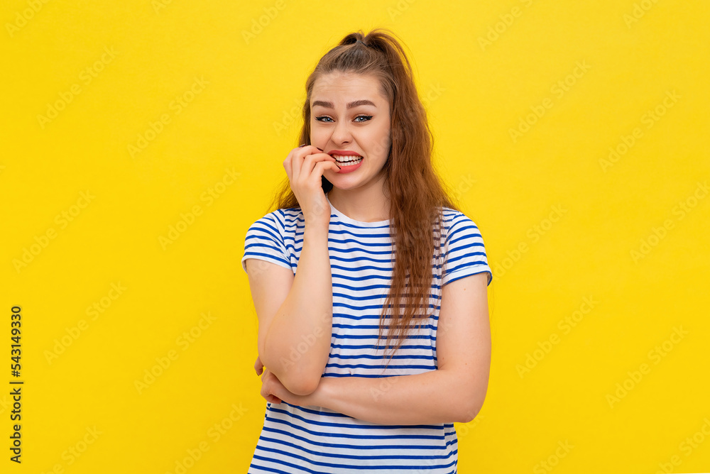 Insecure young woman biting fingernails, looking scared, trembling fear, watching something scary, standing anxious in white-blue striped t shirt over yellow background Insecure young woman biting fingernails, looking scared, trembling fear, watching something scary, standing anxious in white-blue striped t shirt over yellow background
