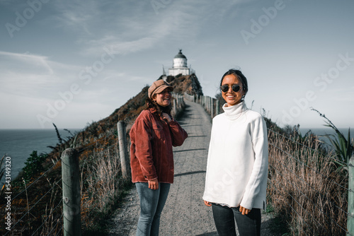 two caucasian young women standing dressed in white pullover red jacket black and blue pants having fun laughing on dirt road leading to mountain lighthouse, nugget point, new zealand photo