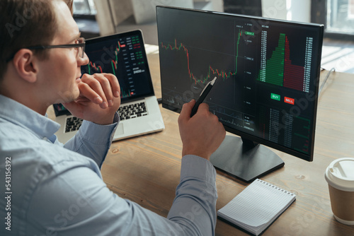 Concentrated businessman trader looking at pc monitor with diagram of stock crypto currency market, pointing with pen at screen, touching chin analyzing tendency of money flow, inventing new strategy