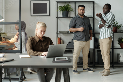 Two intercultural men in casualwear bullying blond Caucasian woman with laptop sitting by desk in front of them and networking photo