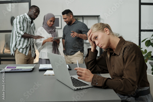 Young tired businesswoman trying to concentrate during work over project while group of multicultural colleagues bullying her