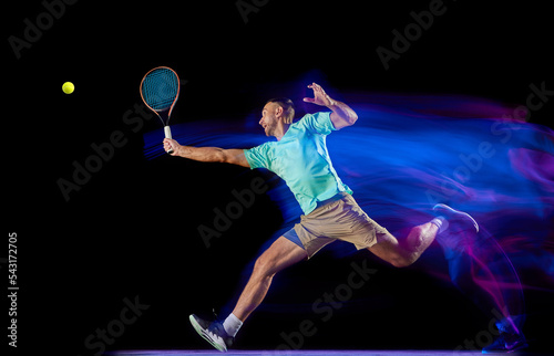 Movements in neon glow. Active male tennis player in sportwear playing tennis isolated over dark background in mixed purple neon light. © master1305