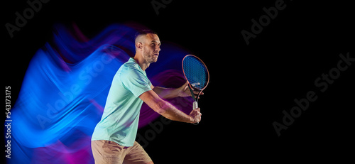 Movements in neon glow. Active male tennis player in sportwear playing tennis isolated over dark background in mixed purple neon light.